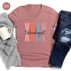 May include: A heather mauve V-neck t-shirt with the text "XRAY technologist" in black lettering. The letters in "XRAY" are stacked vertically and are colored pink, orange, blue, and pink.