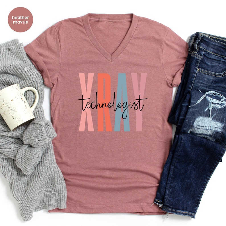 May include: A heather mauve V-neck t-shirt with the text "XRAY technologist" in black lettering. The letters in "XRAY" are stacked vertically and are colored pink, orange, blue, and pink.