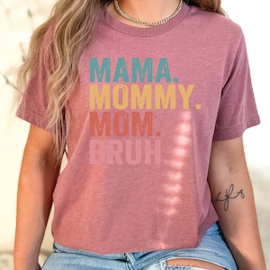May include: A mauve colored t-shirt with the text "MAMA. MOMMY. MOM. BRUH." printed in a retro style font with different colors for each word.