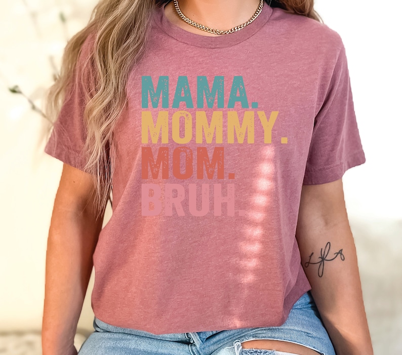 May include: A mauve colored t-shirt with the text "MAMA. MOMMY. MOM. BRUH." printed in a retro style font with different colors for each word.