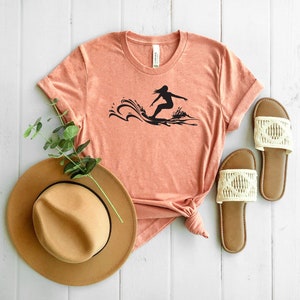May include: A coral colored t-shirt with a black silhouette of a person surfing on a wave. The t-shirt is laying on a white wooden surface with a brown hat and two white sandals.