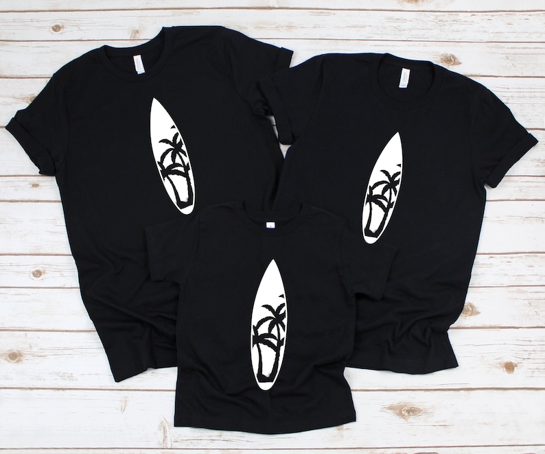 May include: Three black t-shirts with a white silhouette of a surfboard and palm trees. The shirts are arranged in a pyramid shape, with the largest shirt at the top and the smallest shirt at the bottom.