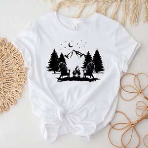 Pu&ograve; includere: Maglietta bianca con un design a silhouette nera di due sedie di fronte a un fal&ograve; davanti a una catena montuosa con alberi e una luna crescente nel cielo.