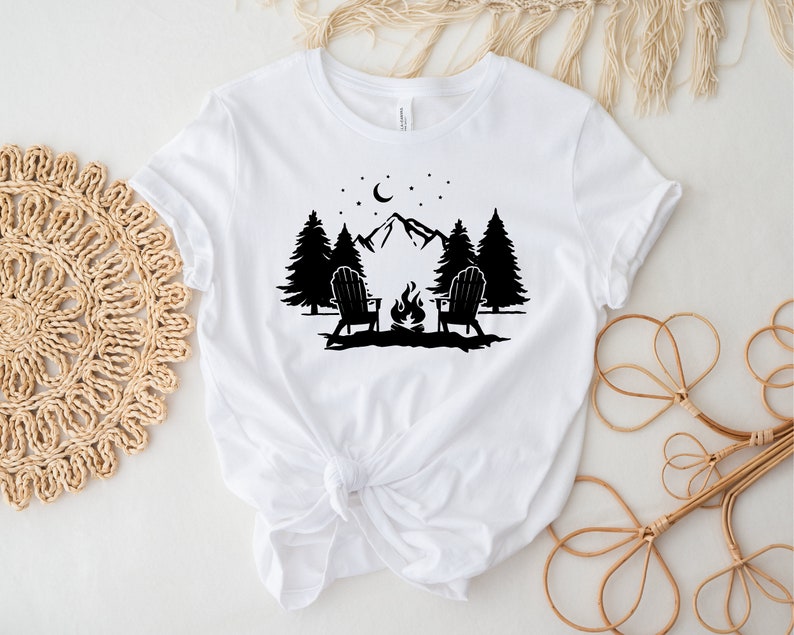 Pu&ograve; includere: Maglietta bianca con un design a silhouette nera di due sedie di fronte a un fal&ograve; davanti a una catena montuosa con alberi e una luna crescente nel cielo.