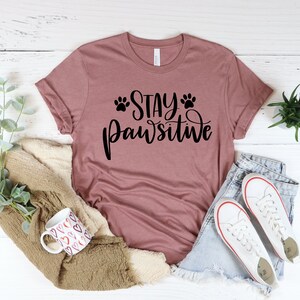 May include: A mauve t-shirt with the text "Stay Pawsitive" in black lettering with paw prints on either side of the word "Stay".