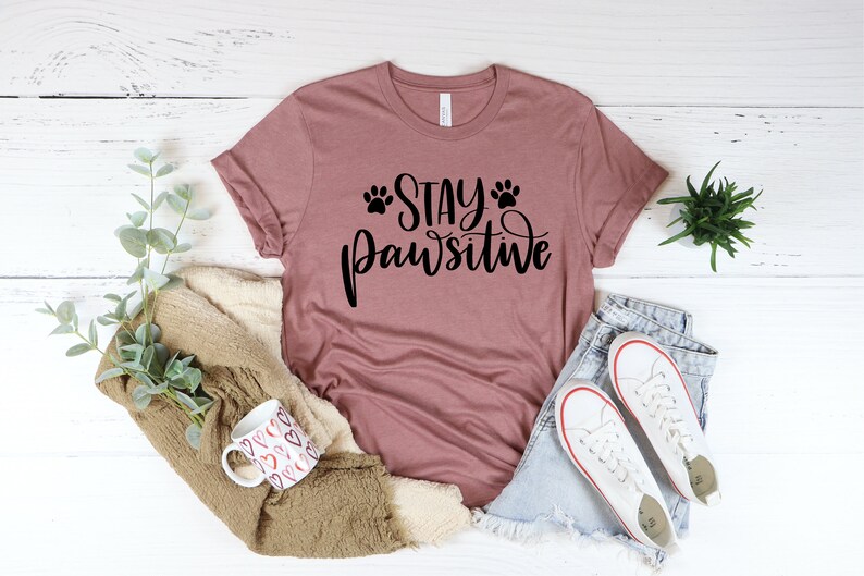 May include: A mauve t-shirt with the text "Stay Pawsitive" in black lettering with paw prints on either side of the word "Stay".