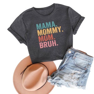 May include: A dark gray t-shirt with the text "MAMA. MOMMY. MOM. BRUH." in colorful retro-style lettering.
