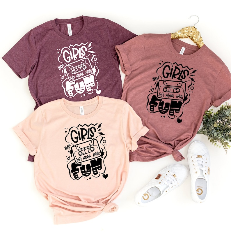 K&ouml;nnte beinhalten: Drei T-Shirts mit einem grafischen Design eines Kassettenbandes mit dem Text "80's Girls Just Wanna Have Fun" in Schwarz auf wei&szlig;em Hintergrund. Die T-Shirts sind in verschiedenen Farben: Rotbraun, Rosa und Hellrosa.