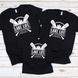 Puede incluir: Camisetas negras con un gr&aacute;fico blanco que dice "LAKE LIFE" encima de "MAKING LIFELONG MEMORIES". El dise&ntilde;o incluye remos cruzados, &aacute;rboles y una l&iacute;nea de agua. Se muestran tres camisetas de diferentes tama&ntilde;os.