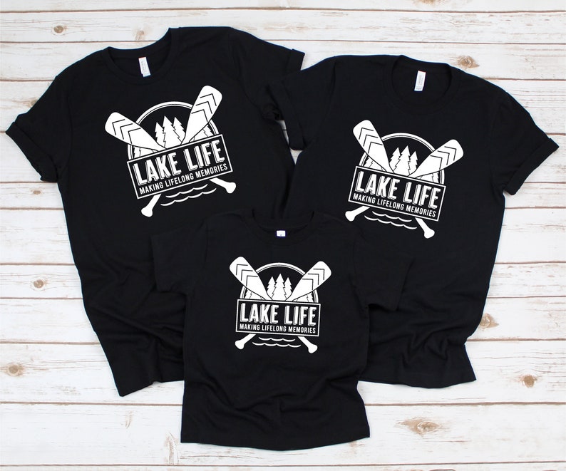 Puede incluir: Camisetas negras con un gr&aacute;fico blanco que dice "LAKE LIFE" encima de "MAKING LIFELONG MEMORIES". El dise&ntilde;o incluye remos cruzados, &aacute;rboles y una l&iacute;nea de agua. Se muestran tres camisetas de diferentes tama&ntilde;os.