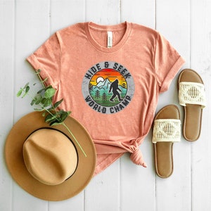 May include: A peach colored t-shirt with a vintage style graphic of a Bigfoot in a forest with the text "Hide & Seek World Champ". The shirt is laid out on a white wood surface with a brown hat and two white sandals.