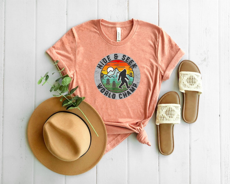 May include: A peach colored t-shirt with a vintage style graphic of a Bigfoot in a forest with the text "Hide & Seek World Champ". The shirt is laid out on a white wood surface with a brown hat and two white sandals.