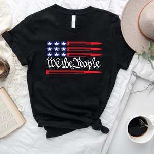 May include: Black t-shirt with a red, white, and blue American flag design. The flag has bullets in place of the red stripes. The text "We the People" is written in a stylized font below the flag.