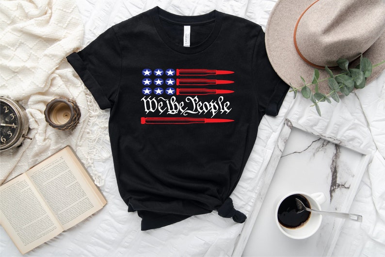 May include: Black t-shirt with a red, white, and blue American flag design. The flag has bullets in place of the red stripes. The text "We the People" is written in a stylized font below the flag.