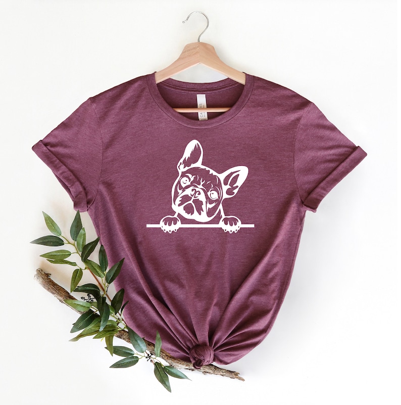 Puede incluir: Una camiseta de color granate con un gr&aacute;fico blanco de un bulldog franc&eacute;s asom&aacute;ndose por encima de una superficie. El bulldog mira directamente al espectador.