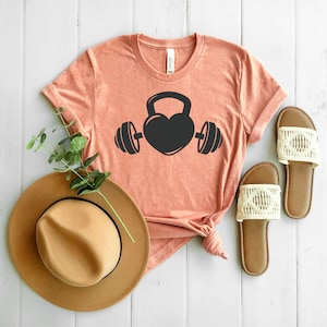 May include: A coral colored t-shirt with a black graphic of a heart shaped kettlebell with a barbell on each side.