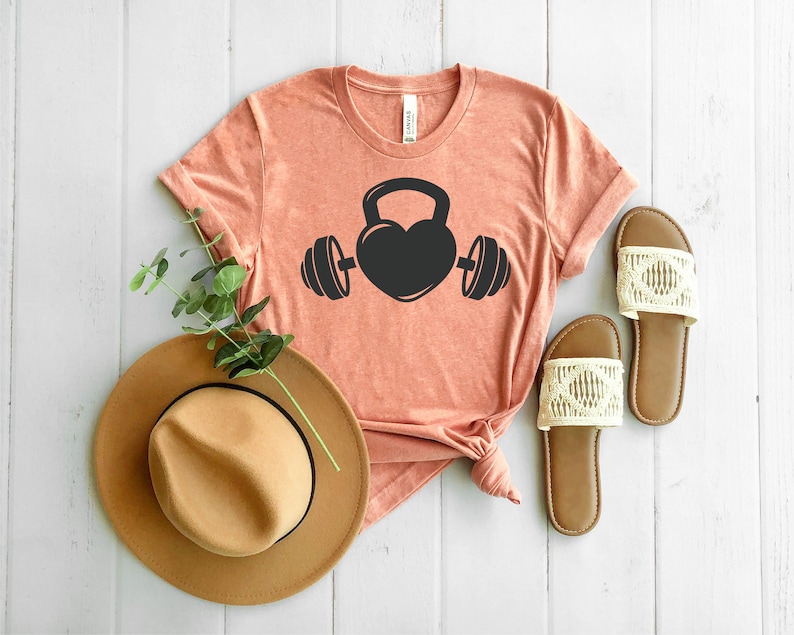 May include: A coral colored t-shirt with a black graphic of a heart shaped kettlebell with a barbell on each side.