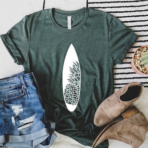 May include: A green t-shirt with a white pineapple design on a surfboard. The shirt is laying on a white and black striped rug with a pair of brown ankle boots and a small potted cactus.