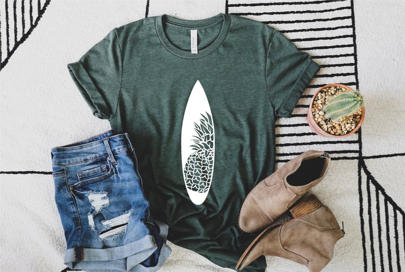 May include: A green t-shirt with a white pineapple design on a surfboard. The shirt is laying on a white and black striped rug with a pair of brown ankle boots and a small potted cactus.