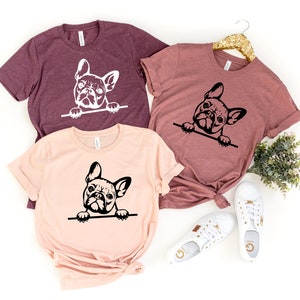 Puede incluir: Tres camisetas con un gr&aacute;fico en blanco y negro de un bulldog franc&eacute;s asom&aacute;ndose por la parte superior de la camiseta. Las camisetas son de diferentes colores: granate, rosa y un tono granate m&aacute;s claro.