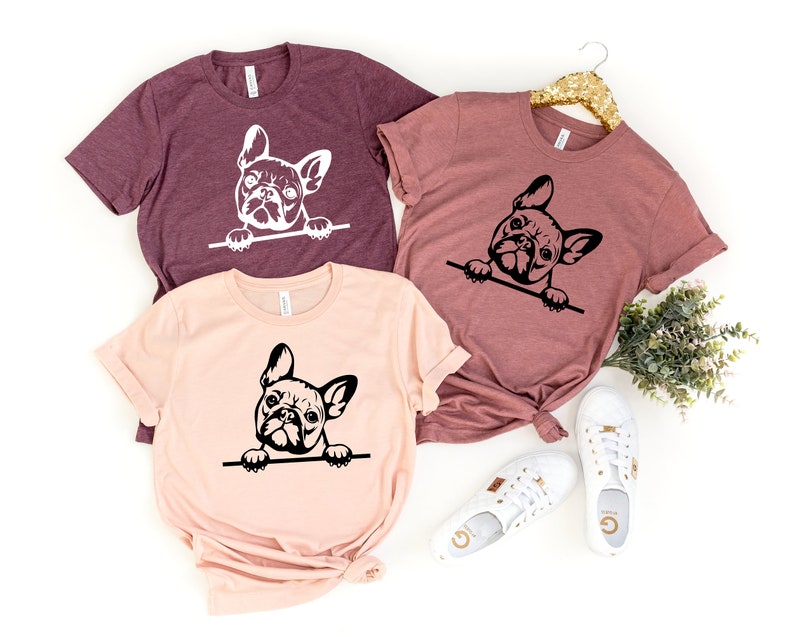 Puede incluir: Tres camisetas con un gr&aacute;fico en blanco y negro de un bulldog franc&eacute;s asom&aacute;ndose por la parte superior de la camiseta. Las camisetas son de diferentes colores: granate, rosa y un tono granate m&aacute;s claro.