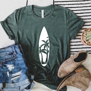 May include: A green t-shirt with a white graphic of a surfboard with palm trees on it. The shirt is laying on a white and black striped rug with a pair of brown boots and a cactus in a terracotta pot.