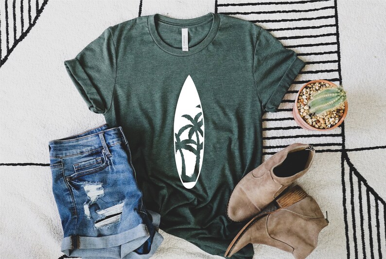 May include: A green t-shirt with a white graphic of a surfboard with palm trees on it. The shirt is laying on a white and black striped rug with a pair of brown boots and a cactus in a terracotta pot.