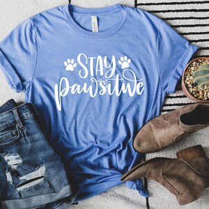 May include: A blue t-shirt with the text "Stay Pawsitive" in white lettering with paw prints on either side of the word "Stay".