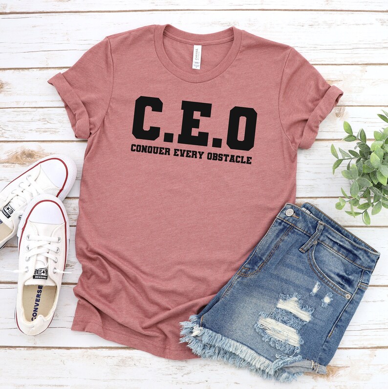 May include: A pink t-shirt with the text "C.E.O. CONQUER EVERY OBSTACLE" in black lettering. The shirt is paired with denim shorts.