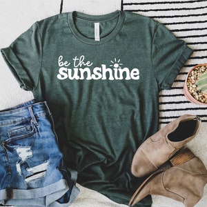 May include: A green t-shirt with the text "be the sunshine" in white lettering. The shirt is laid out on a white and black striped rug with a pair of brown ankle boots and a potted cactus.