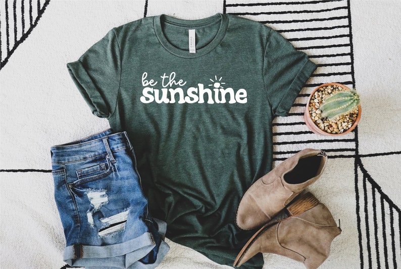 May include: A green t-shirt with the text "be the sunshine" in white lettering. The shirt is laid out on a white and black striped rug with a pair of brown ankle boots and a potted cactus.