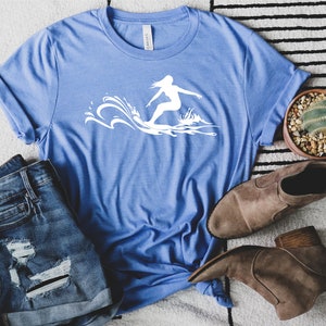 May include: A blue t-shirt with a white silhouette of a person surfing on a wave.