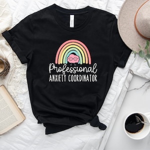 May include: Black t-shirt featuring a colorful rainbow design with a brain wearing glasses. The text reads "Professional Anxiety Coordinator" in white script. The shirt is styled on a white surface with a book, clock, and coffee.