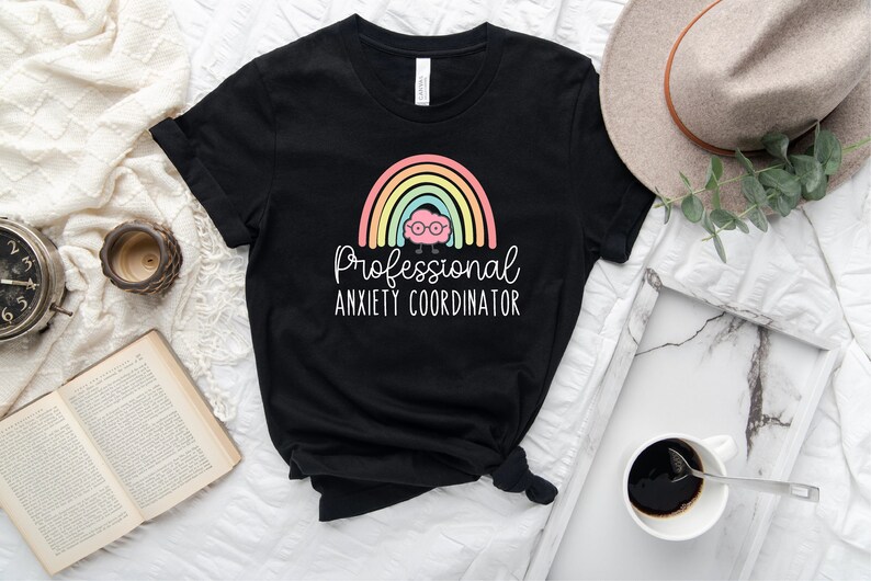 May include: Black t-shirt featuring a colorful rainbow design with a brain wearing glasses. The text reads "Professional Anxiety Coordinator" in white script. The shirt is styled on a white surface with a book, clock, and coffee.