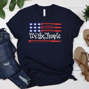 May include: A navy blue t-shirt with a red, white, and blue American flag design. The flag has four red stripes and four white stripes. The stars are white and the text "We the People" is in red. The flag is overlaid with four red bullets.