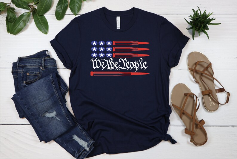 May include: A navy blue t-shirt with a red, white, and blue American flag design. The flag has four red stripes and four white stripes. The stars are white and the text "We the People" is in red. The flag is overlaid with four red bullets.