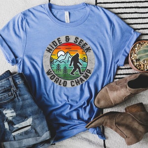 May include: A blue t-shirt with a vintage-style graphic of a Bigfoot in a mountain landscape. The graphic says "Hide & Seek World Champ".