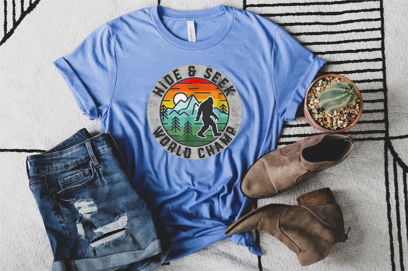 May include: A blue t-shirt with a vintage-style graphic of a Bigfoot in a mountain landscape. The graphic says "Hide & Seek World Champ".