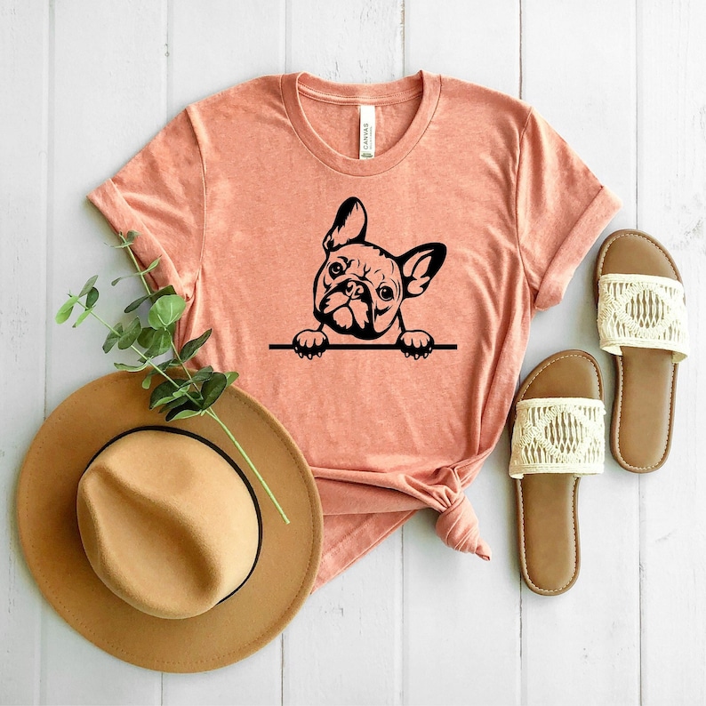 Puede incluir: Una camiseta de color melocot&oacute;n con una silueta negra de un bulldog franc&eacute;s asom&aacute;ndose por la parte superior. La camiseta est&aacute; sobre una superficie blanca con un sombrero marr&oacute;n y unas sandalias blancas.