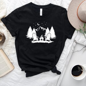Pu&ograve; includere: Maglietta nera con un design grafico bianco di due sedie intorno a un fal&ograve; di fronte a una montagna con alberi e stelle.