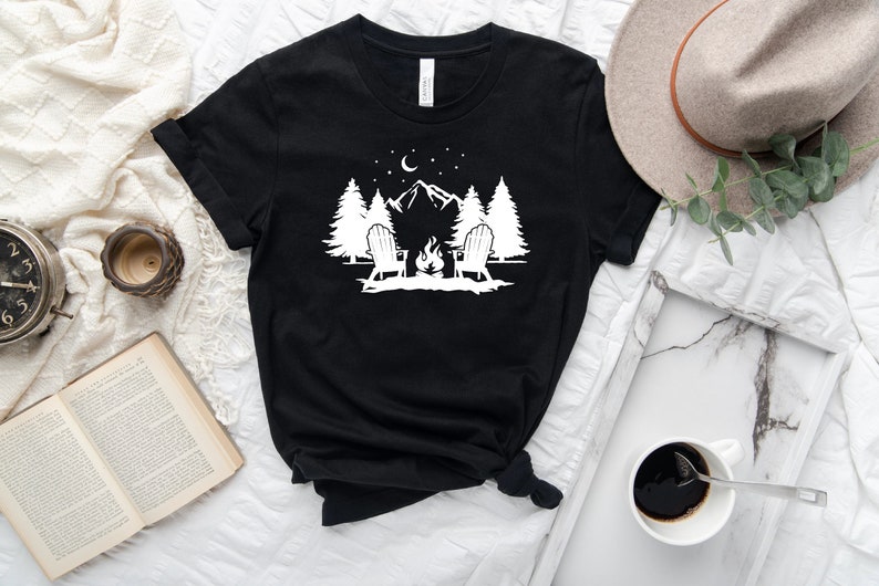 Pu&ograve; includere: Maglietta nera con un design grafico bianco di due sedie intorno a un fal&ograve; di fronte a una montagna con alberi e stelle.