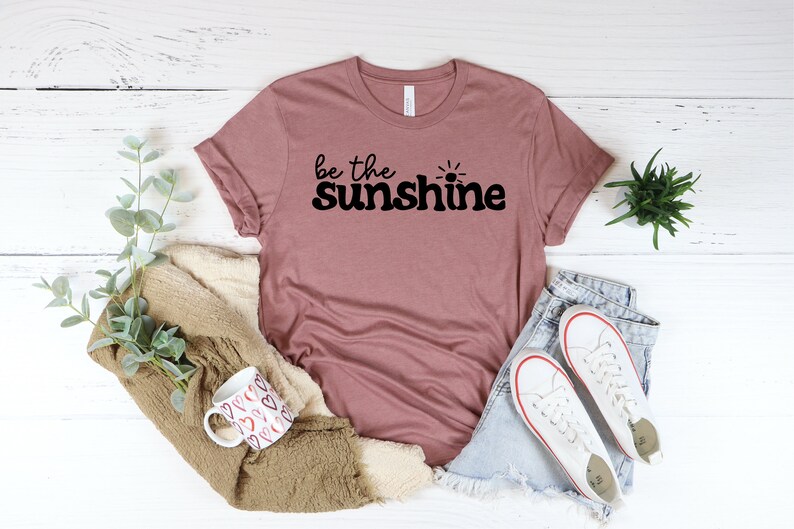 May include: A mauve t-shirt with the text "be the sunshine" in black lettering with a small sun graphic.