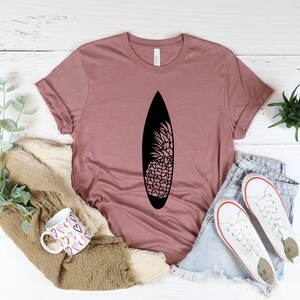 May include: A mauve t-shirt with a black pineapple design on a surfboard. The shirt is laid out on a white wooden surface with a mug, a blanket, and a pair of white sneakers.