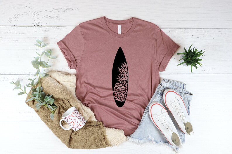May include: A mauve t-shirt with a black pineapple design on a surfboard. The shirt is laid out on a white wooden surface with a mug, a blanket, and a pair of white sneakers.