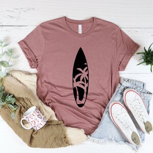 May include: A mauve t-shirt with a black silhouette of a surfboard with palm trees on it. The shirt is laid flat on a white wooden surface with a pair of white sneakers, a mug, and a plant.