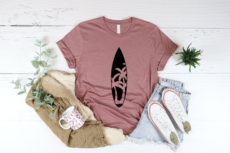 May include: A mauve t-shirt with a black silhouette of a surfboard with palm trees on it. The shirt is laid flat on a white wooden surface with a pair of white sneakers, a mug, and a plant.