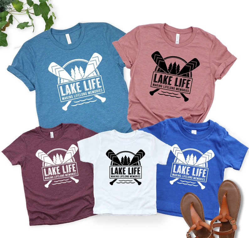 Puede incluir: Varias camisetas de manga corta en varios colores, incluyendo azul, rosa, blanco, burdeos y azul real. Cada camiseta presenta el texto "LAKE LIFE MAKING LIFELONG MEMORIES" con remos cruzados.