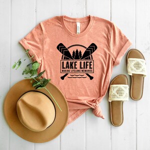 Puede incluir: Una camiseta color melocot&oacute;n con el texto "LAKE LIFE MAKING LIFELONG MEMORIES" en negro. El dise&ntilde;o incluye remos cruzados, &aacute;rboles y una ilustraci&oacute;n de un lago. Tambi&eacute;n se ven un sombrero marr&oacute;n y sandalias con detalles de macram&eacute; color crema.