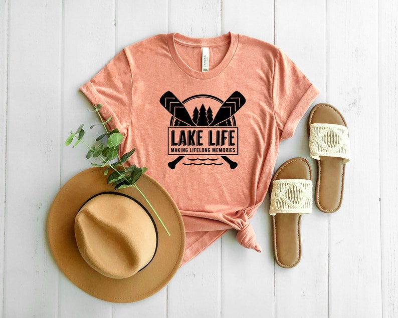Puede incluir: Una camiseta color melocot&oacute;n con el texto "LAKE LIFE MAKING LIFELONG MEMORIES" en negro. El dise&ntilde;o incluye remos cruzados, &aacute;rboles y una ilustraci&oacute;n de un lago. Tambi&eacute;n se ven un sombrero marr&oacute;n y sandalias con detalles de macram&eacute; color crema.