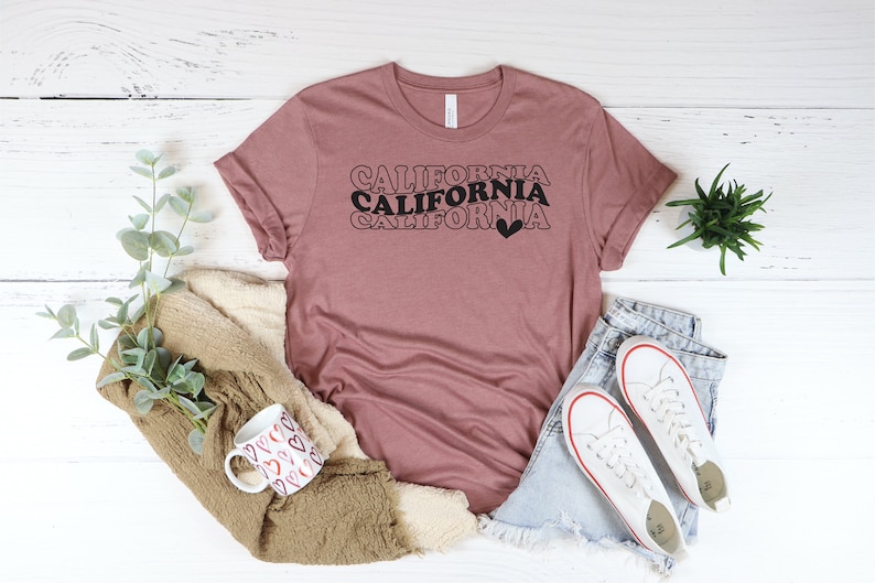 Puede incluir: Una camiseta color malva con el texto "CALIFORNIA" repetido tres veces en un dise&ntilde;o apilado con un peque&ntilde;o coraz&oacute;n al final. El texto est&aacute; en negro con un contorno rosa.